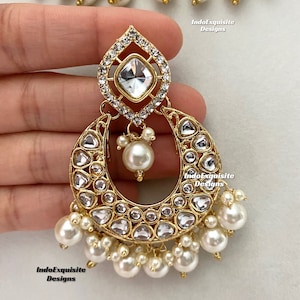 May include: Close-up of a gold-toned earring with a teardrop-shaped top, a crescent-shaped body, and dangling pearls. The earring is adorned with clear, sparkling crystals and a large, square-cut crystal at the top. The jewelry is part of a set.