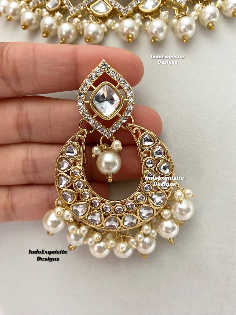 May include: Close-up of a gold-toned earring with a teardrop-shaped top, a crescent-shaped body, and dangling pearls. The earring is adorned with clear, sparkling crystals and a large, square-cut crystal at the top. The jewelry is part of a set.