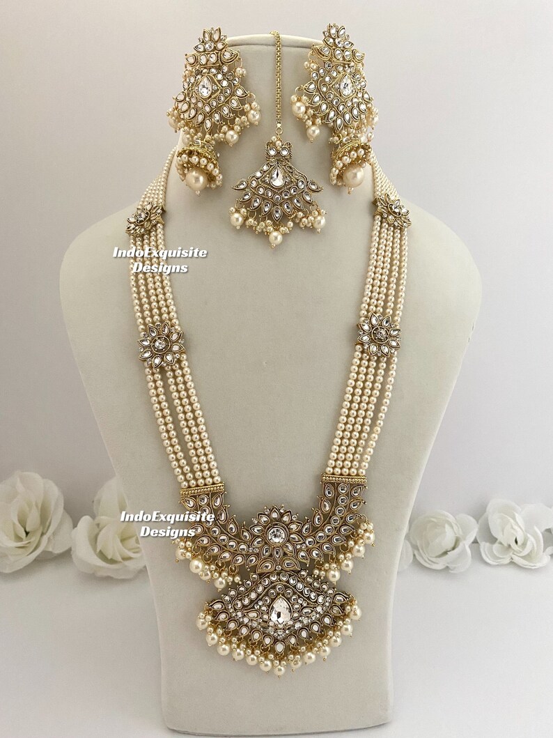 May include: An ornate, gold-toned jewelry set featuring a multi-strand pearl necklace, matching earrings, and a headpiece. The necklace has a large, detailed pendant with clear crystals and pearl accents. The earrings and headpiece mirror the pendant's design.