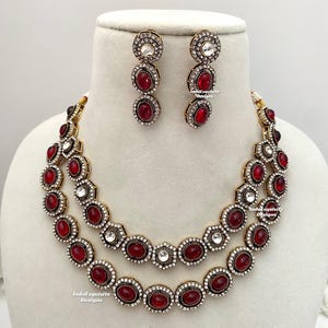 May include: A two-tiered necklace and earrings set with red and clear stones. The necklace has a gold tone finish and features oval-shaped red stones surrounded by clear stones. The earrings are drop earrings with a similar design.