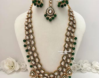 May include: A gold and green Indian bridal necklace set with a long, layered necklace, matching earrings, and a tikka. The necklace features intricate designs with white stones and green beads.