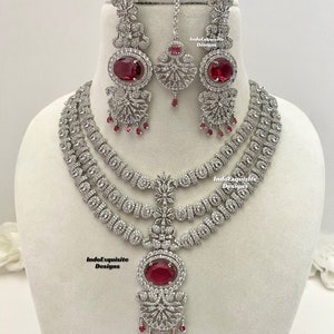 Puede incluir: Un conjunto de joyas que incluye un collar de tres hilos, pendientes y una diadema. El conjunto presenta metal plateado con piedras preciosas claras y rojas. El collar tiene un gran colgante ovalado de piedra roja.