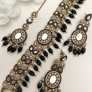 Puede incluir: Un conjunto de joyas de novia indias en oro y negro con un collar choker, pendientes y un maang tikka. El conjunto presenta espejos redondos, cuentas negras y perlas blancas.