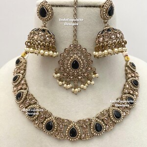 May include: An antique gold-toned jewelry set featuring a necklace, earrings, and a headpiece. The set includes black teardrop-shaped stones, clear stones, and small white pearls. The earrings are bell-shaped. The text "IndoExquisite Designs" is visible.