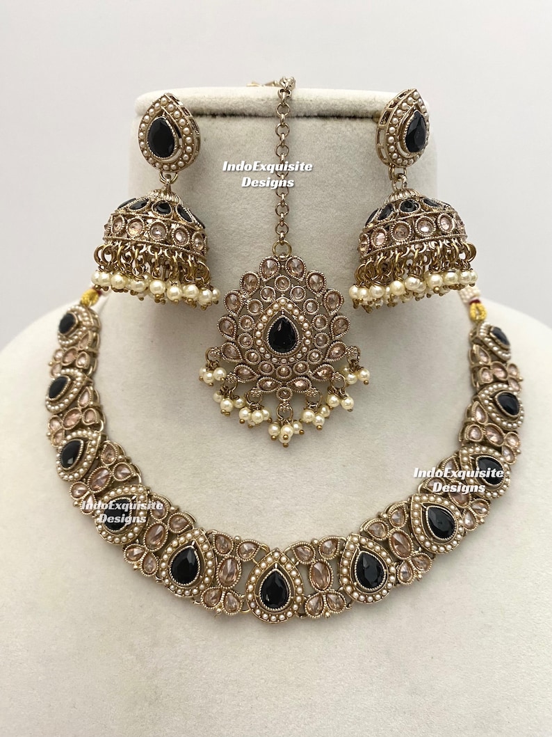 May include: An antique gold-toned jewelry set featuring a necklace, earrings, and a headpiece. The set includes black teardrop-shaped stones, clear stones, and small white pearls. The earrings are bell-shaped. The text "IndoExquisite Designs" is visible.