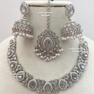 May include: A silver-toned jewelry set featuring a necklace, earrings, and a headpiece. The necklace and earrings have a teardrop design with clear stones and small white pearls. The headpiece has a central teardrop stone and a chain.