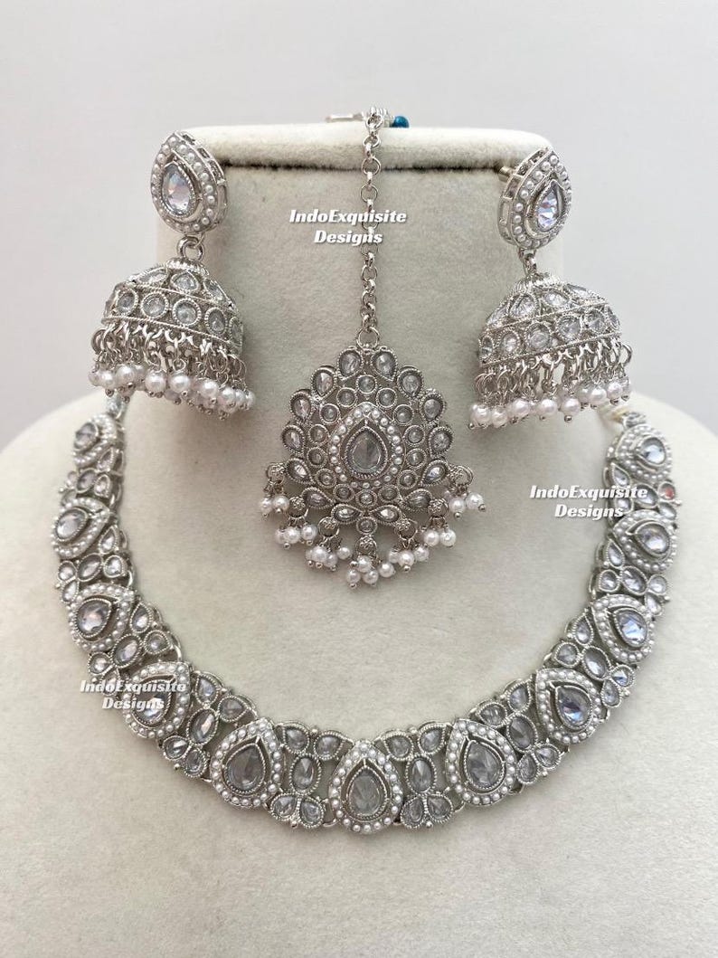 May include: A silver-toned jewelry set featuring a necklace, earrings, and a headpiece. The necklace and earrings have a teardrop design with clear stones and small white pearls. The headpiece has a central teardrop stone and a chain.
