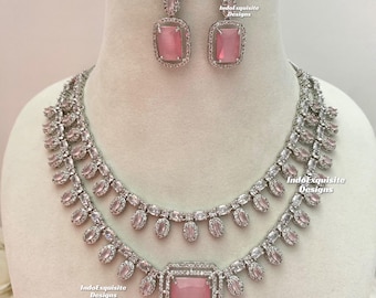 May include: A silver necklace with a pink stone pendant and matching earrings. The necklace has a double layer of oval-shaped stones and a larger square-shaped stone in the center. The earrings have a pink stone and two smaller stones.