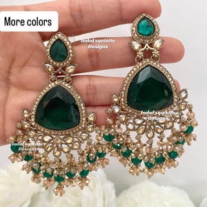 May include: A pair of elaborate gold earrings with large, emerald-green, triangular and teardrop-shaped gemstones. The earrings have smaller clear and green beads and are set against a soft, blurred background. The text "More colours" is visible.