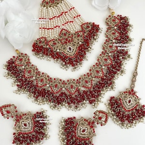 May include: A red and gold Indian bridal jewellery set with pearl accents. The set includes a necklace, earrings, a maang tikka, and a nose ring.