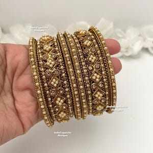 May include: A set of gold-toned bangles with intricate detailing. The bangles are adorned with small, round pearls and decorative patterns. The text "IndoExquisite Designs" is visible on the bangles.