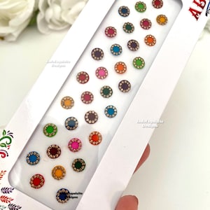 Op de afbeelding: Een vel met 24 kleurrijke ronde bindi-stickers met gouden accenten. De bindi-stickers zijn in rijen op een witte achtergrond gerangschikt. Het vel is gelabeld met "ABR" en "IndoExquisite Designs".