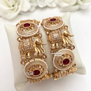 May include: A pair of gold-tone bangles with intricate designs featuring elephants and red gemstones. The bangles are decorated with white pearls and clear rhinestones.