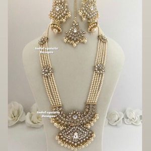 May include: An ornate, cream-colored necklace and earring set with a matching headpiece. The jewelry features multiple strands of small, round pearls, accented with clear crystals and gold-toned metalwork. The set is displayed on a mannequin.