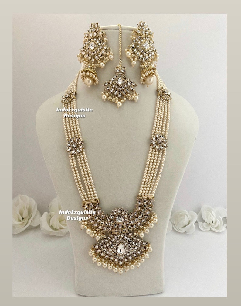 May include: An ornate, cream-colored necklace and earring set with a matching headpiece. The jewelry features multiple strands of small, round pearls, accented with clear crystals and gold-toned metalwork. The set is displayed on a mannequin.