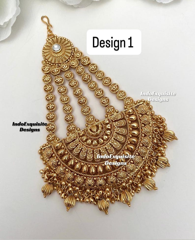 May include: Gold-toned headpiece with intricate floral and beaded details. The design features a central circular element with a clear stone, cascading strands, and a decorative lower section. The text "Design 1" is visible.
