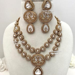 May include: A gold-toned jewelry set featuring a necklace and matching earrings. The necklace has two strands of round, clear stones and a central oval pendant with a floral design. The earrings have a similar design.