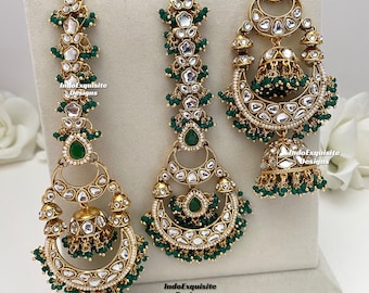 May include: An ornate gold-coloured bridal jewellery set with clear and green detailing. The set includes a headpiece and earrings, all decorated with small, dangling green beads and clear stones. The pieces are displayed on a white stand.