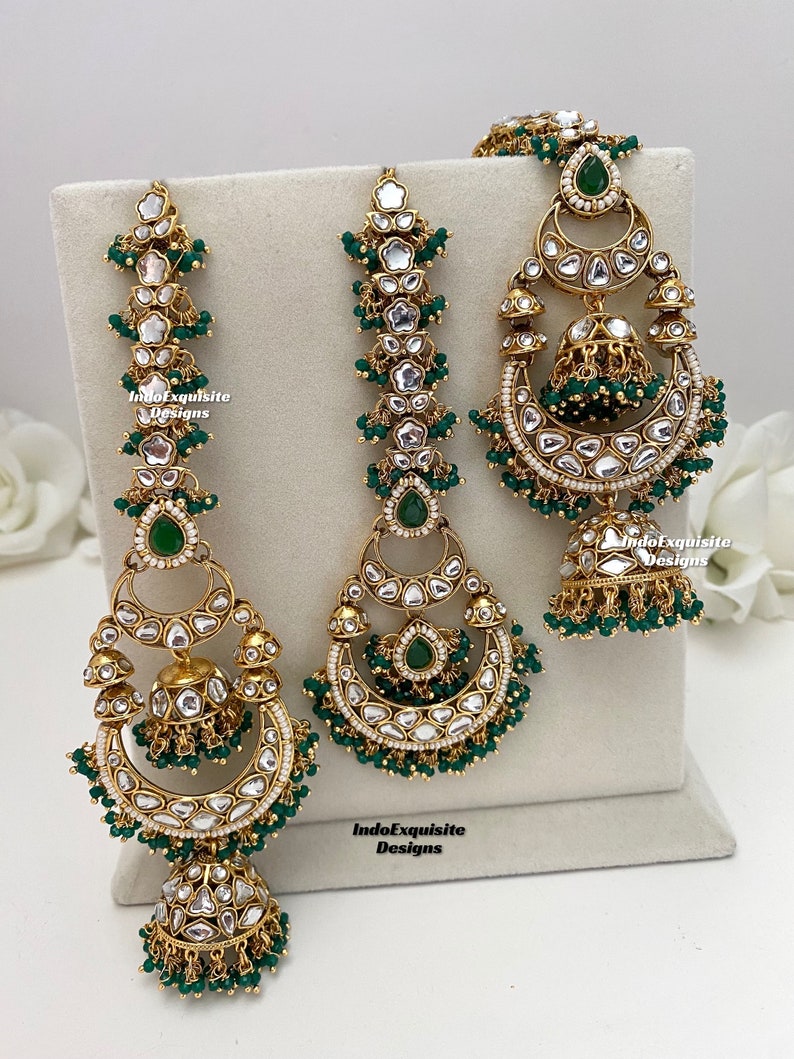 Puede incluir: Un conjunto de joyer&iacute;a en tono dorado que incluye dos adornos verticales largos para el cabello y un par de pendientes a juego. Cada pieza est&aacute; decorada con piedras blancas, detalles en forma de gota verdes y peque&ntilde;as cuentas verdes. Texto IndoExquisite Designs.