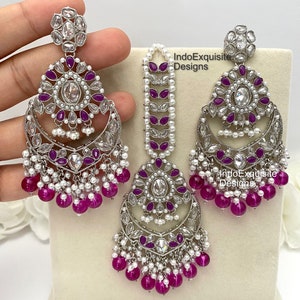 May include: A silver and white jewelry set with pink and white beads. The set includes a pair of earrings and a maang tikka. The earrings are large and have a crescent moon shape with a floral design. The maang tikka is a long, thin piece with a floral design at the top. The set is made of silver and white metal with pink and white beads.