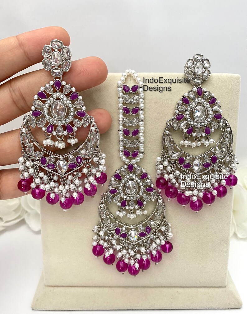 May include: A silver and white jewelry set with pink and white beads. The set includes a pair of earrings and a maang tikka. The earrings are large and have a crescent moon shape with a floral design. The maang tikka is a long, thin piece with a floral design at the top. The set is made of silver and white metal with pink and white beads.
