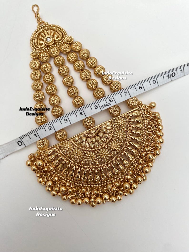 May include: A gold-toned headpiece featuring a semi-circular pendant with intricate floral engravings and dangling beads, connected to four vertical chains of small circular discs. A measuring tape shows the piece is approximately 7.5 inches long.