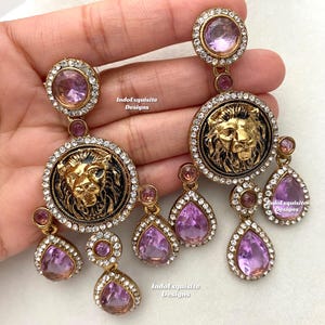 May include: A pair of gold-tone earrings with a lion head design and dangling purple crystals. The earrings are adorned with clear rhinestones.