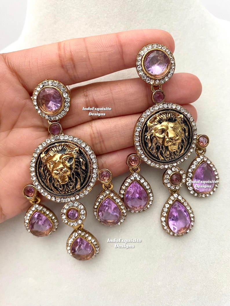 May include: A pair of gold-tone earrings with a lion head design and dangling purple crystals. The earrings are adorned with clear rhinestones.