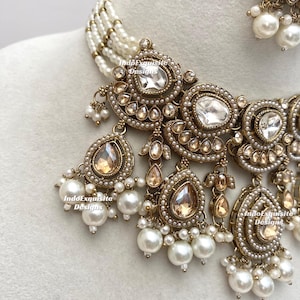 May include: Close-up of an ornate necklace and earring set. The jewelry features a gold-toned metal with clear and champagne-colored stones, and white pearls. The necklace has multiple strands of small pearls. The brand name "IndoExquisite Designs" is visible.