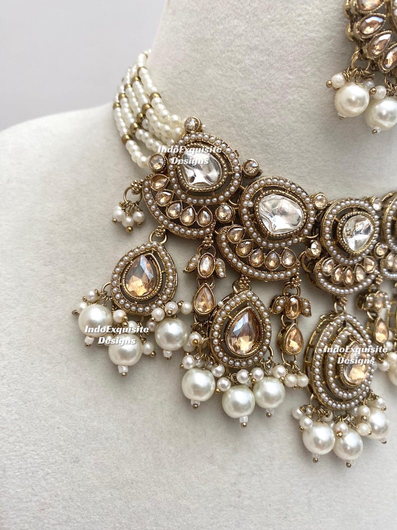May include: Close-up of an ornate necklace and earring set. The jewelry features a gold-toned metal with clear and champagne-colored stones, and white pearls. The necklace has multiple strands of small pearls. The brand name "IndoExquisite Designs" is visible.