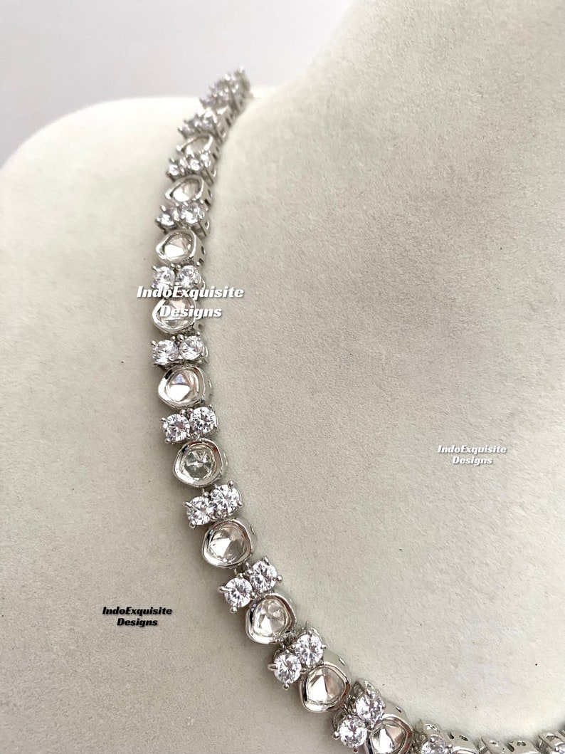 May include: A silver necklace with a row of clear, round stones. The stones are set in a silver setting and are connected by a silver chain. The necklace is on a white background.