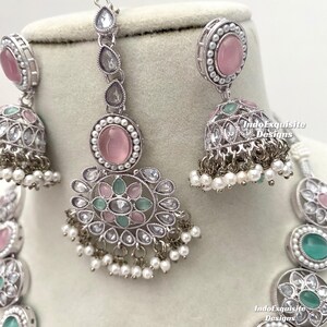 Op de afbeelding: Een zilveren ketting met een ontwerp van roze en groene stenen. De ketting heeft een hanger met een roze steen en een groene steen. De ketting heeft ook een paar oorbellen met roze stenen.