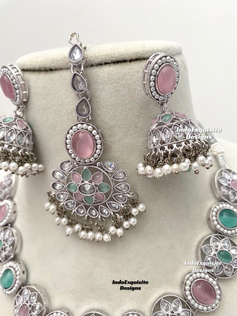 Op de afbeelding: Een zilveren ketting met een ontwerp van roze en groene stenen. De ketting heeft een hanger met een roze steen en een groene steen. De ketting heeft ook een paar oorbellen met roze stenen.