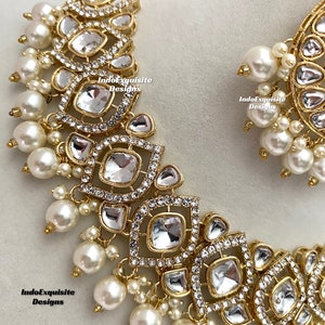 May include: A gold-toned necklace and earring set featuring clear, square-cut stones and small, round, white pearls. The necklace and earrings have a detailed, ornate design with the text "IndoExquisite Designs".