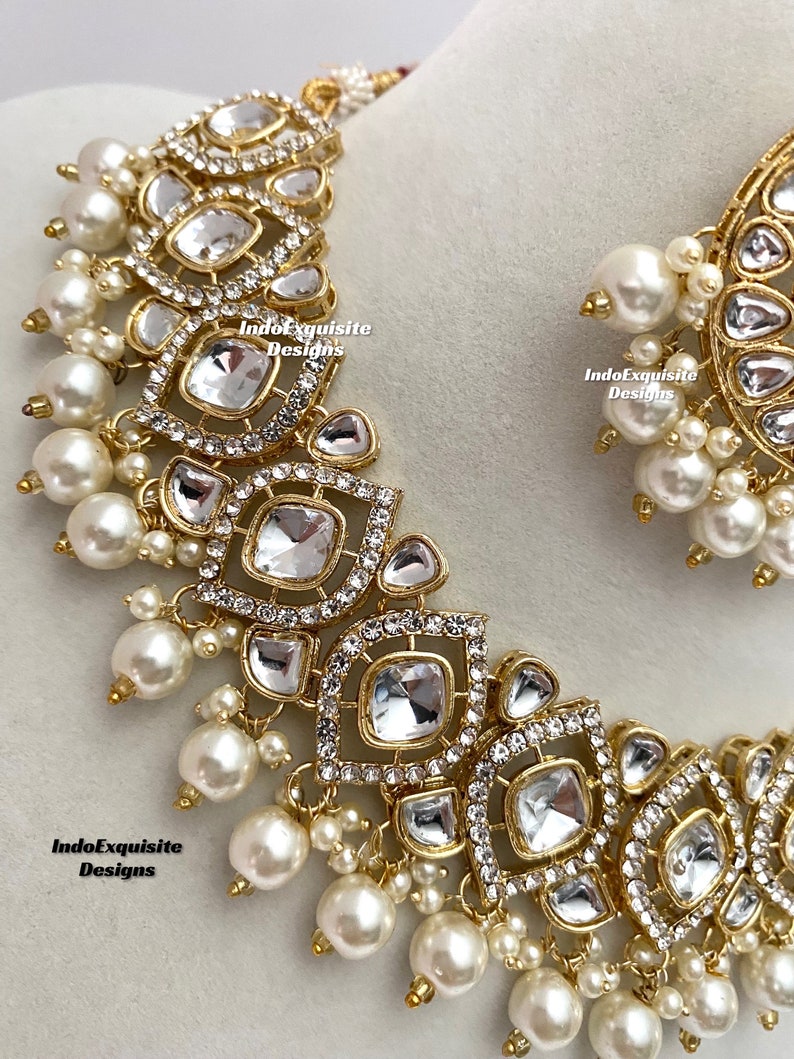 May include: A gold-toned necklace and earring set featuring clear, square-cut stones and small, round, white pearls. The necklace and earrings have a detailed, ornate design with the text "IndoExquisite Designs".