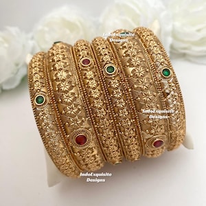 May include: Gold bangles with detailed floral designs and embedded gemstones. The bangles have red and green gemstones. The image includes the text "IndoExquisite Designs".