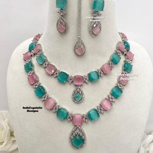May include: A two-tiered silver necklace with a teardrop pendant. The necklace features alternating pink and teal square-shaped stones. Matching earrings with teal and pink stones are also included. The text "IndoExquisite Designs" appears on the image.