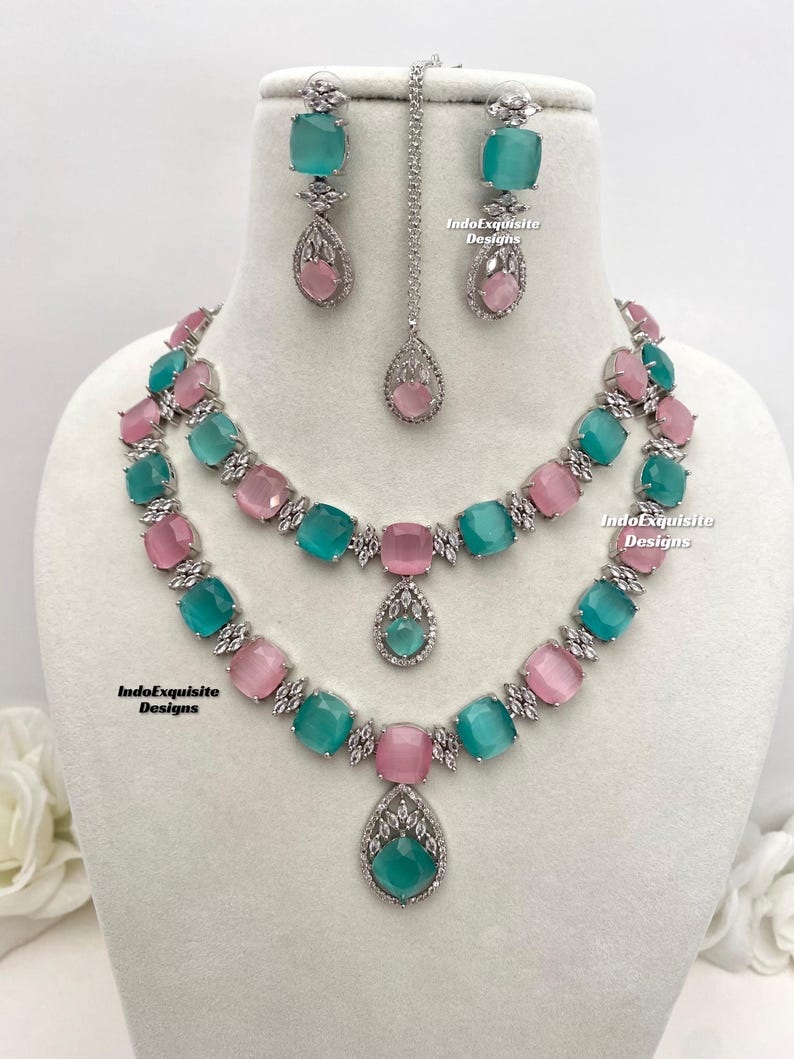 May include: A two-tiered silver necklace with a teardrop pendant. The necklace features alternating pink and teal square-shaped stones. Matching earrings with teal and pink stones are also included. The text "IndoExquisite Designs" appears on the image.