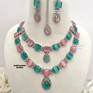 May include: A two-tiered necklace set with a matching pair of earrings and a pendant. The necklace features alternating light pink and teal blue square-shaped stones with clear rhinestones. The earrings and pendant feature a teardrop-shaped teal blue stone with clear rhinestones. The set is made of silver-toned metal. IndoExquisite Designs is printed on the image.