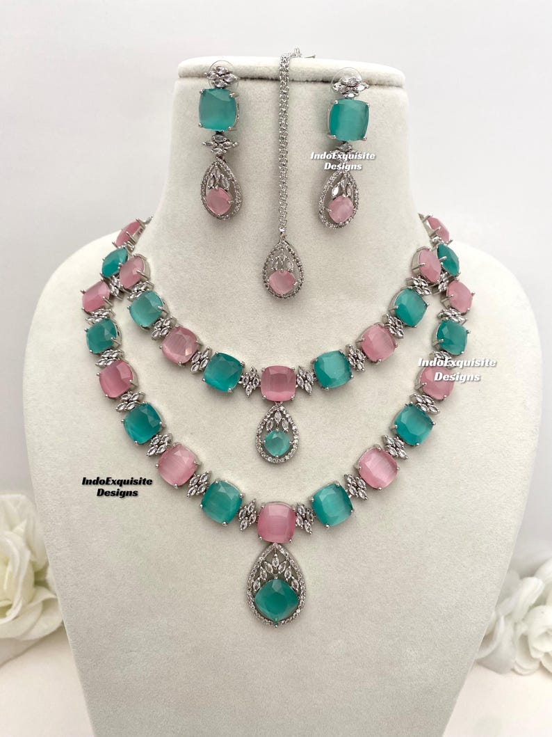May include: A two-tiered necklace set with a matching pair of earrings and a pendant. The necklace features alternating light pink and teal blue square-shaped stones with clear rhinestones. The earrings and pendant feature a teardrop-shaped teal blue stone with clear rhinestones. The set is made of silver-toned metal. IndoExquisite Designs is printed on the image.