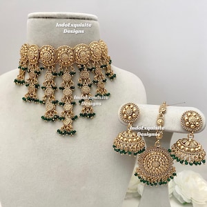 May include: Gold-toned jewelry set featuring a choker necklace and matching earrings. The necklace and earrings have intricate floral designs and dangling green beads. The set is displayed on a mannequin head and stand.