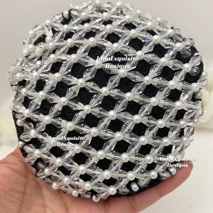 Könnte beinhalten: Ein schwarzes, rundes Haaraccessoire, verziert mit einem Gitter aus klaren, facettierten Perlen und kleinen weißen Perlen. Das Accessoire wird in einer Hand gehalten, wodurch das komplizierte Design und die Textur hervorgehoben werden. Die Worte "IndoExquisite Designs" sind auf dem Accessoire aufgedruckt.