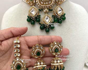 Puede incluir: Un conjunto de joyería en tono dorado que incluye un collar tipo gargantilla, dos pendientes jhumka y un maang tikka. Cada pieza está adornada con perlas blancas, piedras transparentes y detalles verdes en forma de lágrima. El texto IndoExquisite Designs es visible.