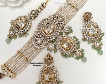 May include: A traditional Indian bridal jewellery set featuring a multi-strand pearl choker necklace, a large statement pendant, and matching earrings. The set is adorned with sparkling clear stones and intricate gold detailing. The earrings feature a teardrop shape with a large central stone and smaller stones cascading down the sides.