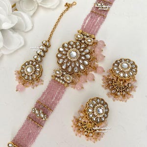 Puede incluir: Un collar choker de cuentas rosas con un gran colgante dorado con diseño floral y un par de aretes a juego más pequeños. El collar y los aretes están adornados con piedras blancas y cuentas rosas.