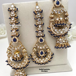 Puede incluir: Un conjunto de joyer&iacute;a en tono dorado que incluye dos pendientes largos y un maang tikka central, adornados con piedras azules en forma de l&aacute;grima, cristales transparentes y peque&ntilde;as cuentas azules colgantes. Incluye el texto IndoExquisite Designs.