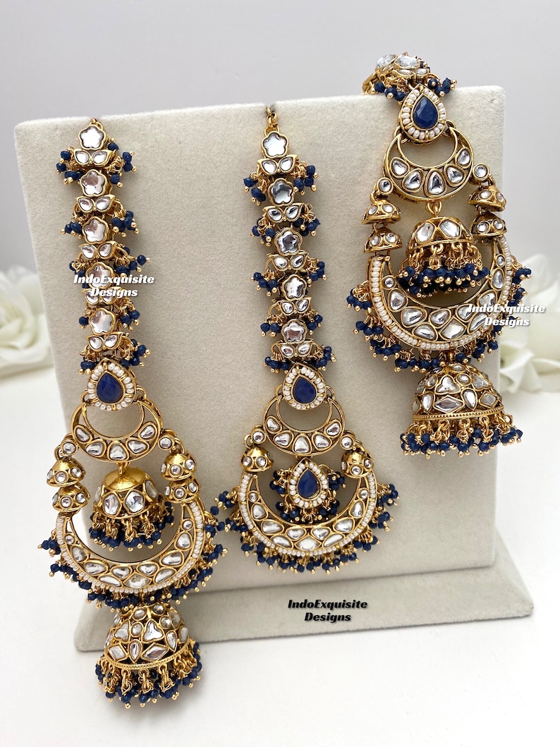 Puede incluir: Un conjunto de joyer&iacute;a en tono dorado que incluye dos pendientes largos y un maang tikka central, adornados con piedras azules en forma de l&aacute;grima, cristales transparentes y peque&ntilde;as cuentas azules colgantes. Incluye el texto IndoExquisite Designs.