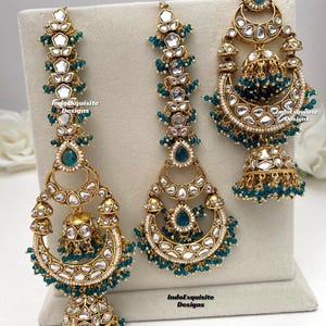 Puede incluir: Un conjunto de joyer&iacute;a en tono dorado que incluye dos adornos largos para el cabello y un pendiente jhumka. Cada pieza est&aacute; decorada con piedras blancas, detalles en forma de gota color verde azulado y peque&ntilde;as cuentas. El texto IndoExquisite Designs es visible.