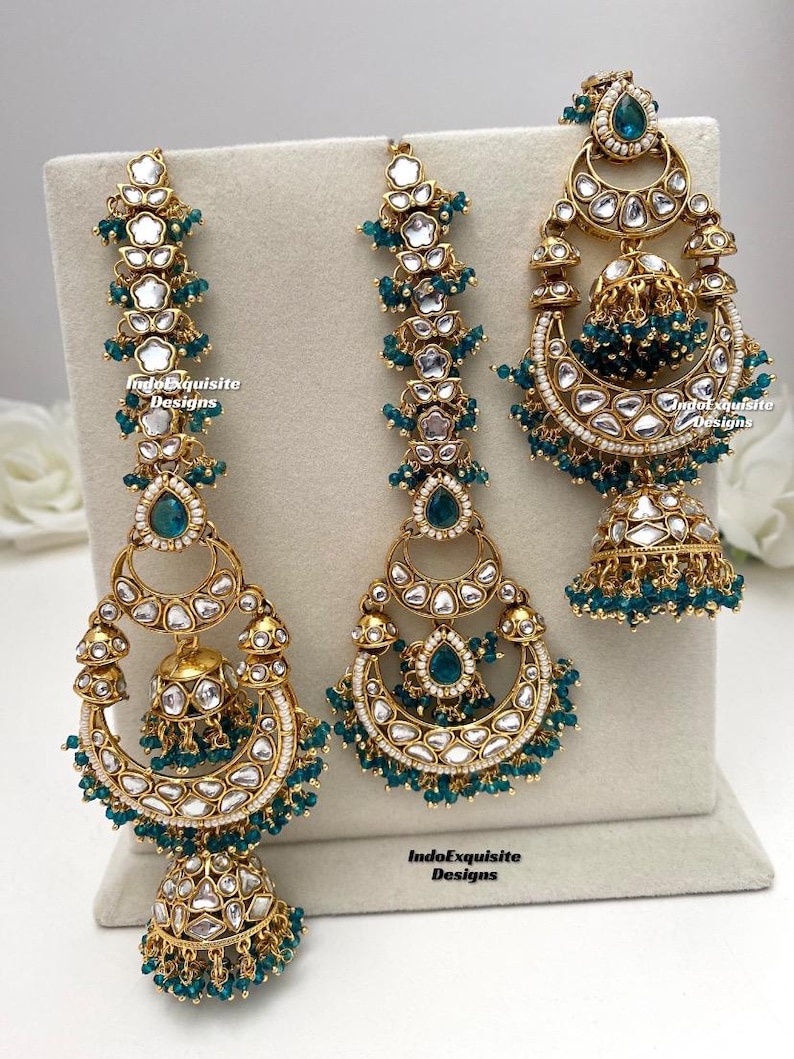 Puede incluir: Un conjunto de joyer&iacute;a en tono dorado que incluye dos adornos largos para el cabello y un pendiente jhumka. Cada pieza est&aacute; decorada con piedras blancas, detalles en forma de gota color verde azulado y peque&ntilde;as cuentas. El texto IndoExquisite Designs es visible.