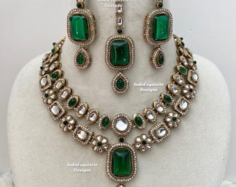Puede incluir: Un conjunto de joyería que incluye un collar de doble capa, dos pendientes colgantes y un maang tikka. Cada pieza está hecha de metal dorado con piedras rectangulares verde esmeralda, piedras claras y motivos florales. El texto dice IndoExquisite Designs.